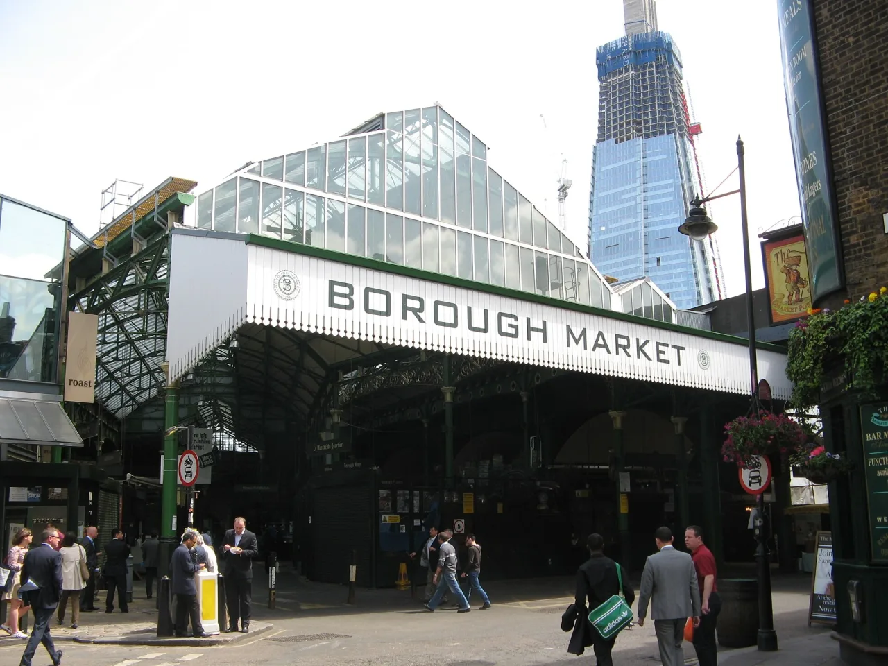 The green iron and glass entrance of Borough Market in London on a bright day, with pedestrians passing and a tall glass tower behind.
