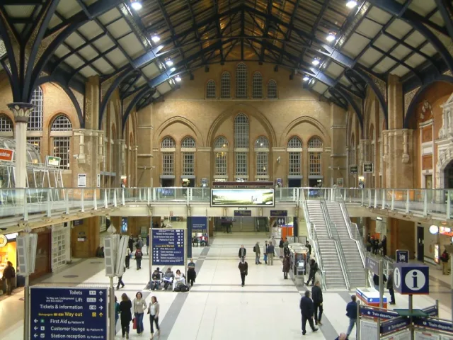 Passengers crossing the main concourse of a London railway terminus, under a high brick hall with arched windows and an iron roof.