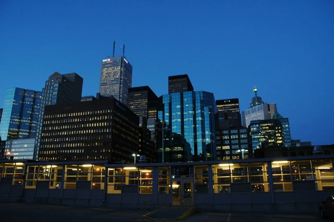 Downtown Toronto office towers lit against a deep blue dusk sky, seen over a low glass-walled building in the foreground.