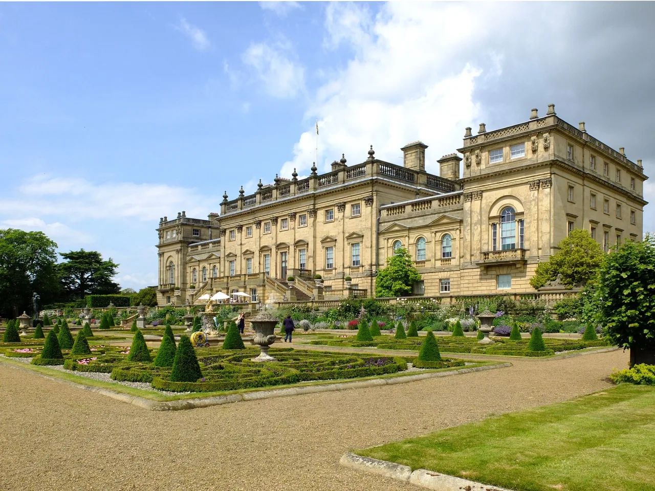 A large honey-coloured stately home seen from its terrace, with clipped topiary and formal flower beds in the foreground