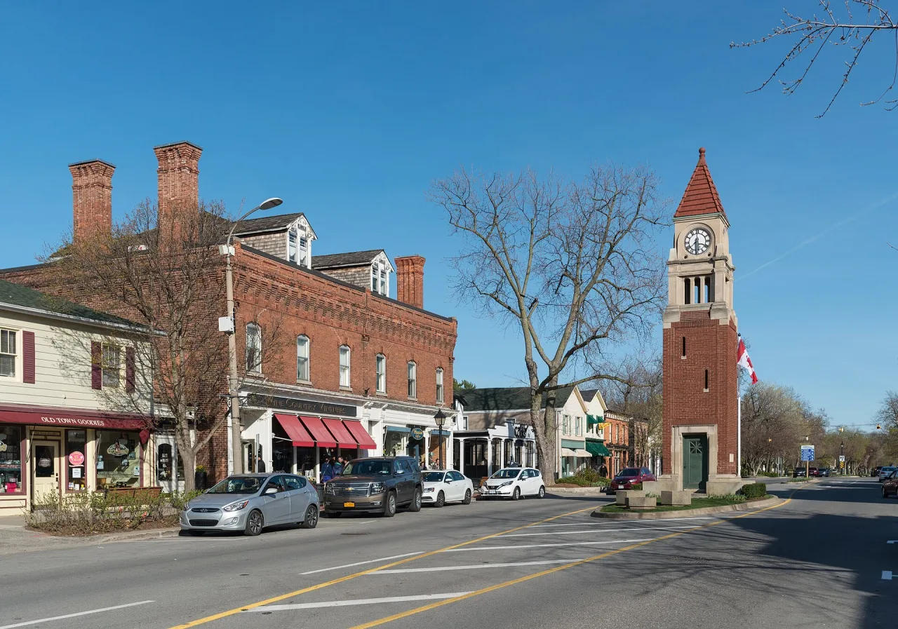 A main street of brick shopfronts with awnings, parked cars and a red-roofed brick clock tower under a clear blue sky.