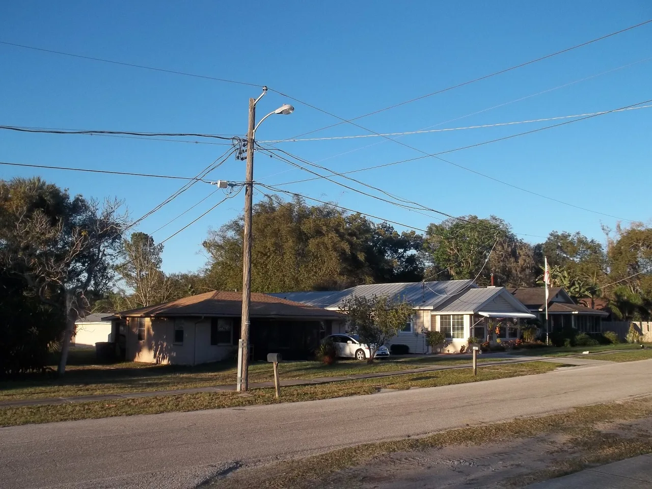 Low single-storey houses with metal roofs along a quiet road, a utility pole with wires in front and trees behind, under a clear blue sky.