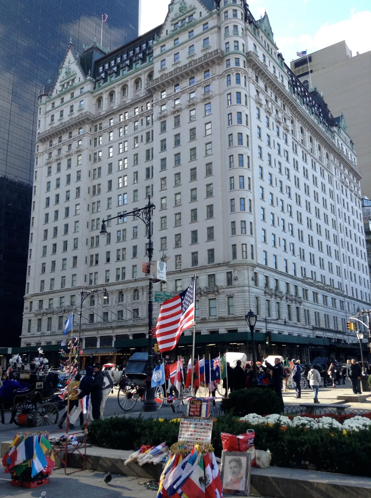 The tall pale stone Plaza Hotel seen from the street, with a large American flag, horse carriages and flag sellers in front.