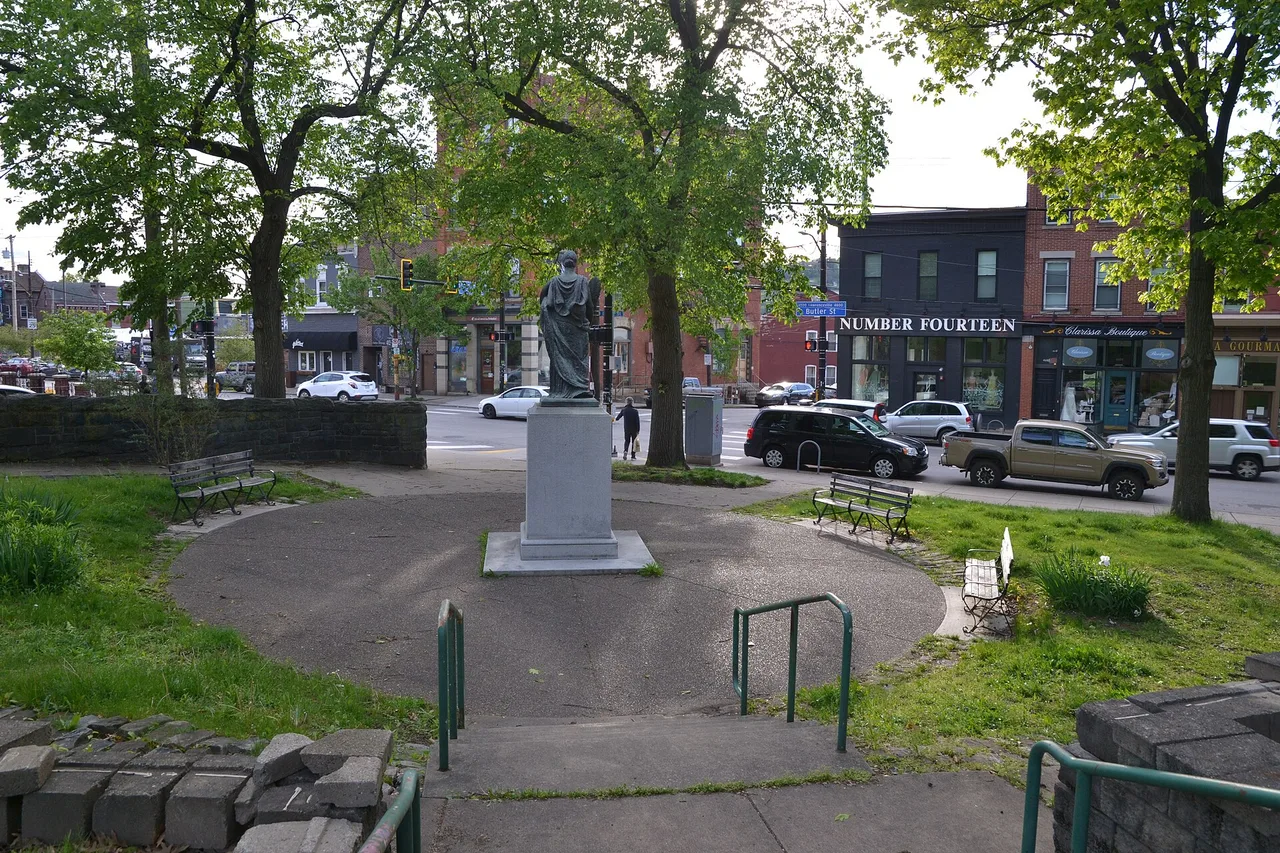 A small green park with a statue on a stone plinth, seen from behind, looking across a street of brick shopfronts and parked cars on a spring day.