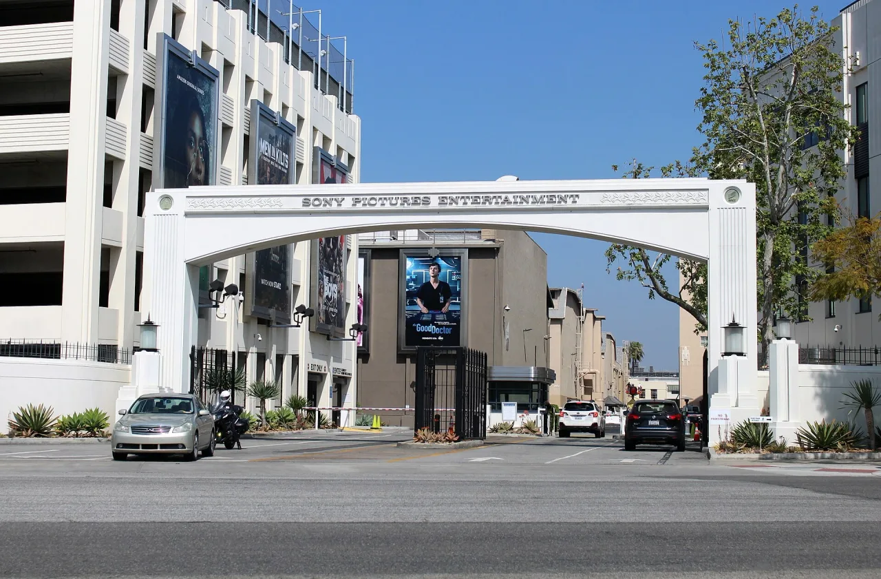 White arched studio gate across a road in sunshine, with large advertising banners on the buildings beside it.