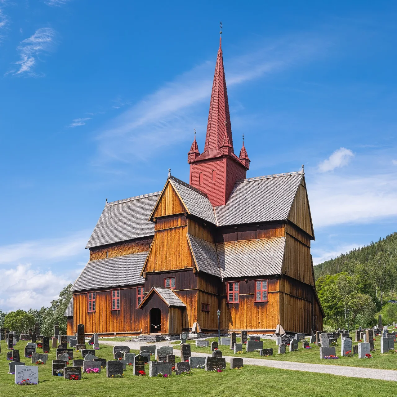 A tall dark-timber stave church with a red spire and grey shingle roofs stands in a green churchyard of headstones under a bright blue sky.