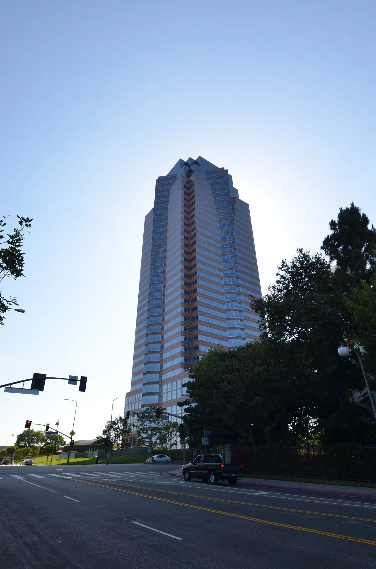 A tall pink-and-blue glass office tower with a stepped crown rising above trees and a road under a clear sky.