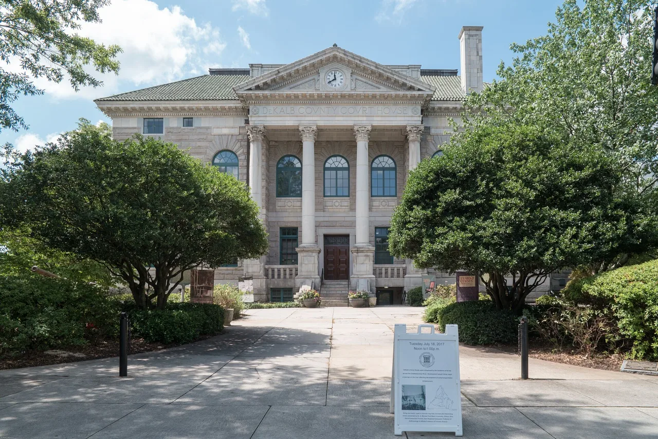 Stone courthouse with a columned front and clock in its pediment, seen from a paved path lined with trees on a sunny day.