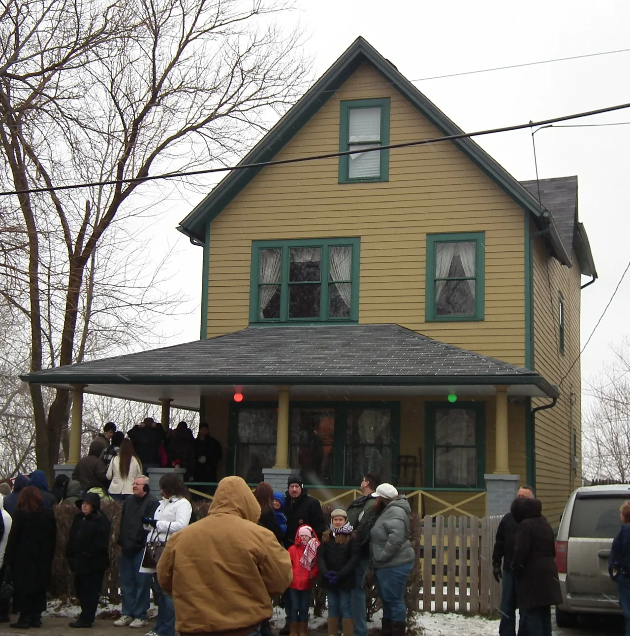 Two-storey yellow clapboard house with green trim and a deep front porch, with a crowd of visitors in winter coats gathered on the pavement.