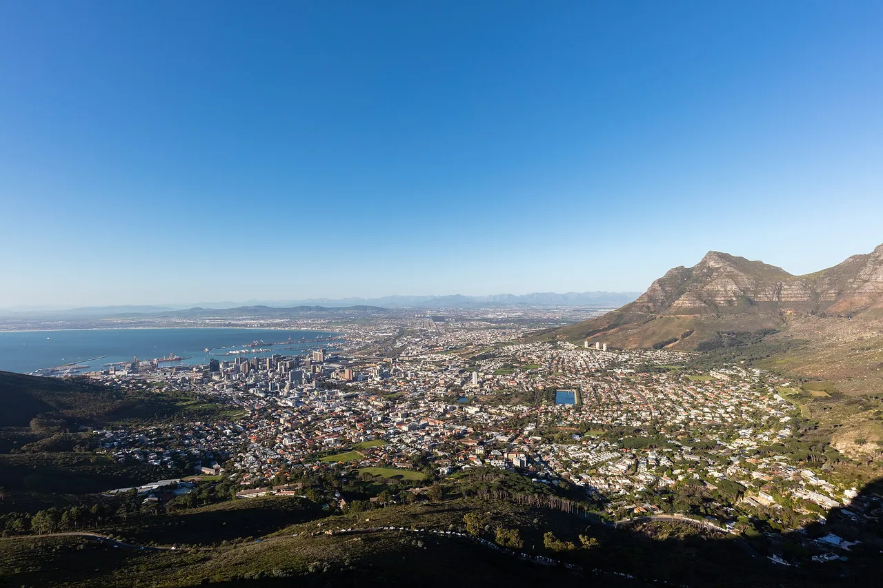 A wide view over Cape Town from a hillside on a clear day, with the harbour on the left, city blocks and suburbs below and a steep mountain on the right.