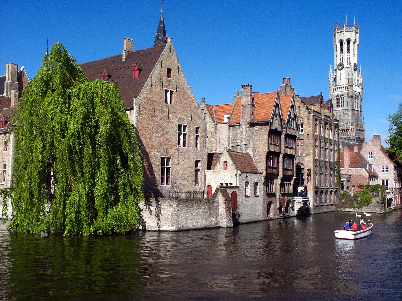 Brick gabled houses beside a canal under a blue sky, a weeping willow on the left, a small boat on the water and the belfry tower rising behind.