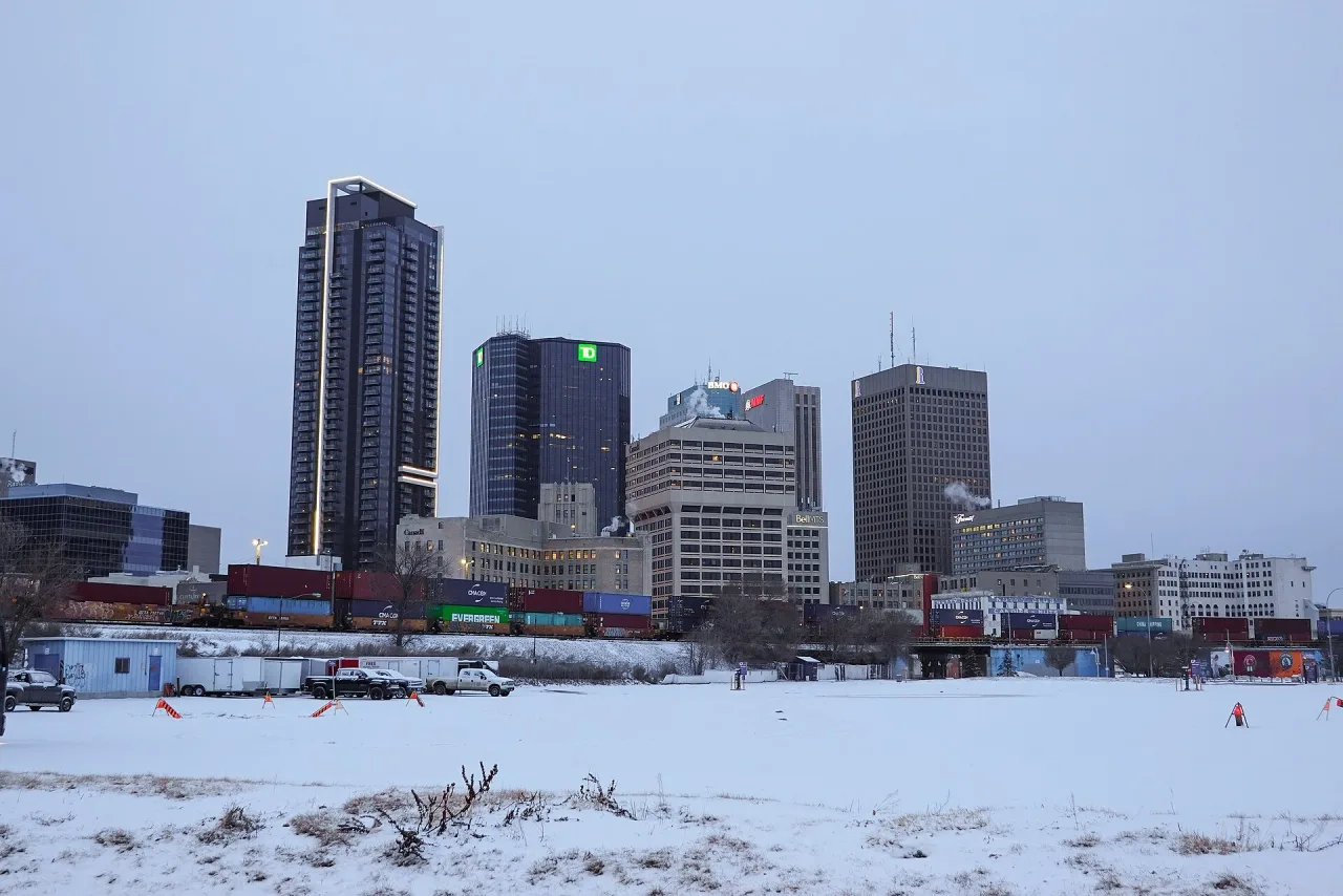 Downtown Winnipeg towers under a grey winter sky, seen across a snow-covered field with a freight train passing in front of the buildings.