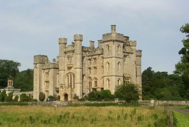 Pale stone castle with turrets and battlements seen across a grassy field, with trees behind and a cloudy sky.