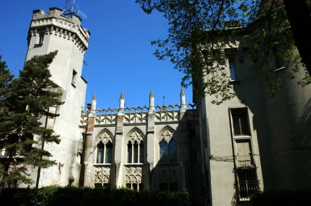 Pale Gothic Revival castle with a crenellated corner tower and pointed arched windows, seen from below against a blue sky