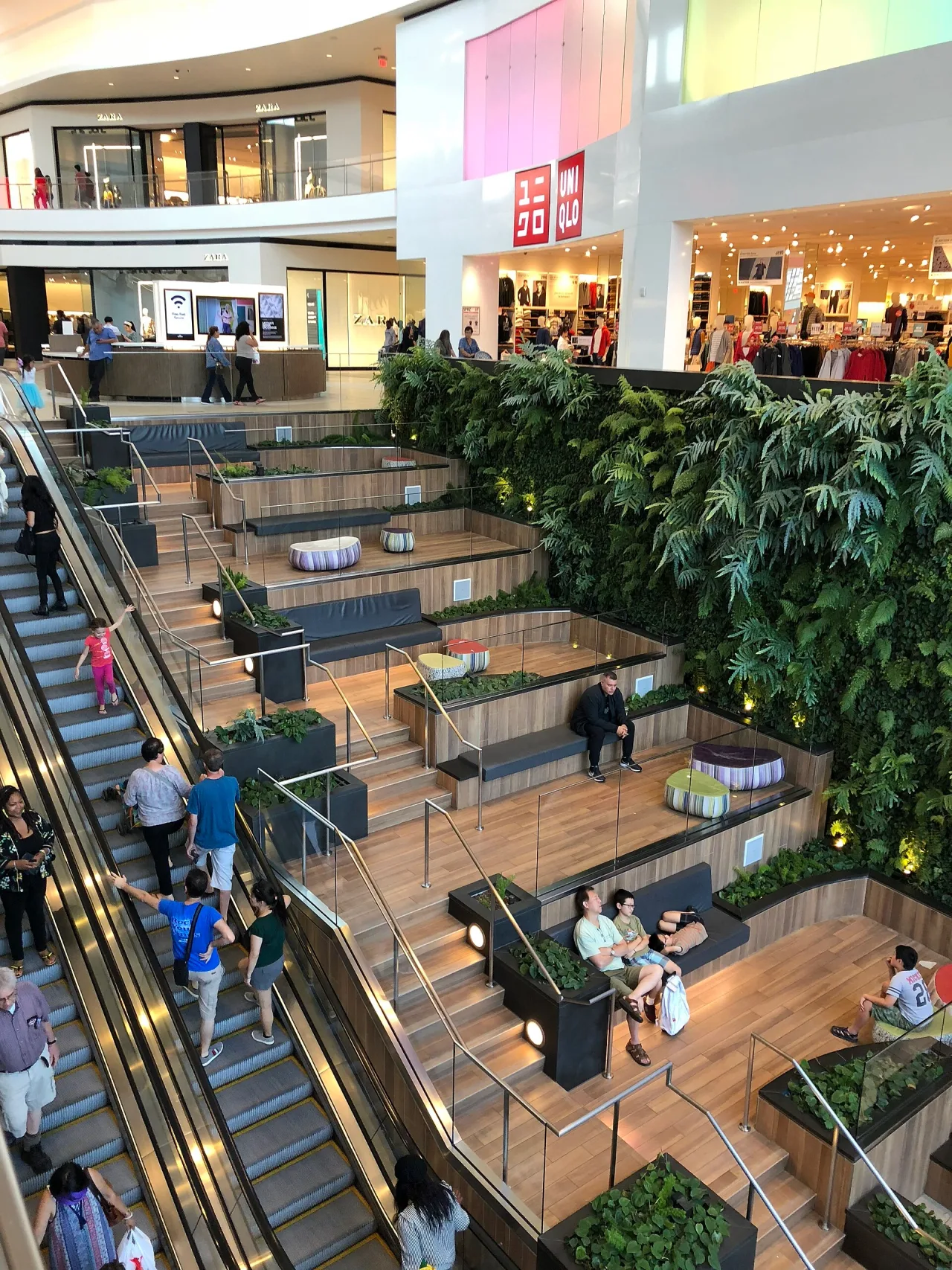 Looking down into a multi-storey shopping centre atrium with escalators, wooden terraced seating, a green planted wall and shoppers on several levels.