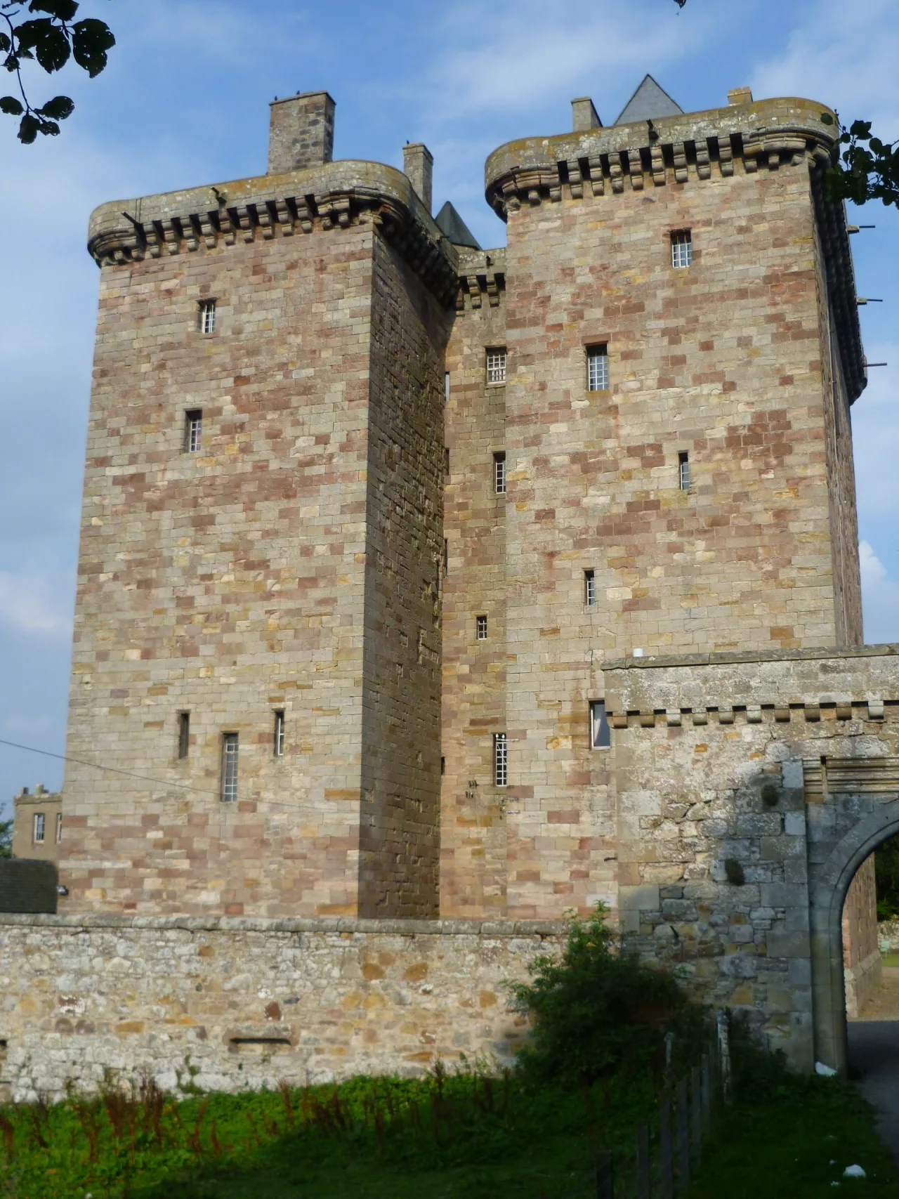 The tall sandstone twin towers of Borthwick Castle rising above a low stone wall and arched gateway under a blue sky.