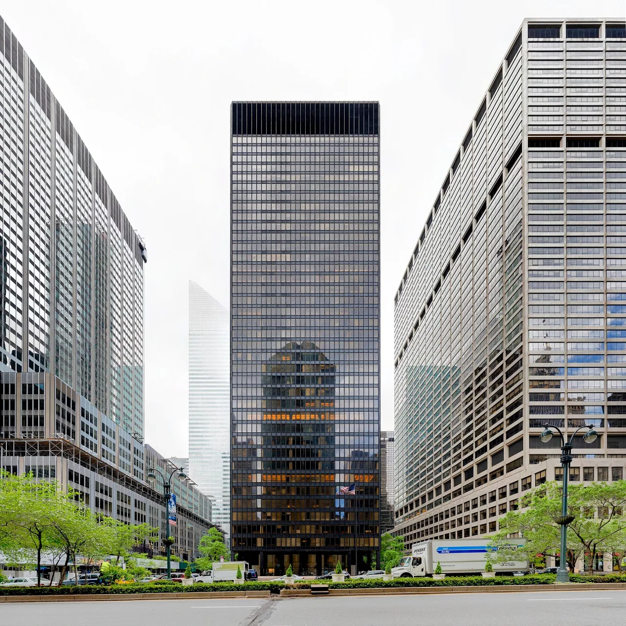 Tall dark glass and bronze office tower seen head-on from across Park Avenue, flanked by lighter towers, with trees and a street lamp in the foreground.