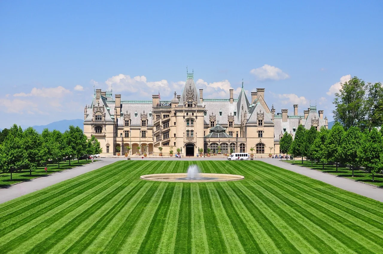 Front of a huge French Renaissance style mansion across a striped green lawn with a round fountain, under a blue sky.
