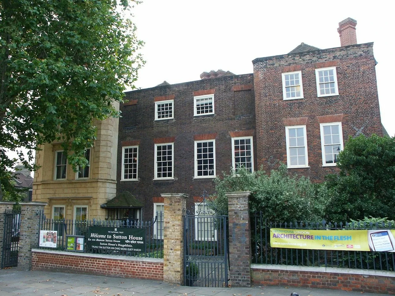 Front of a Tudor-era brick manor house in east London behind iron gates, with a tree on the left and a welcome sign.