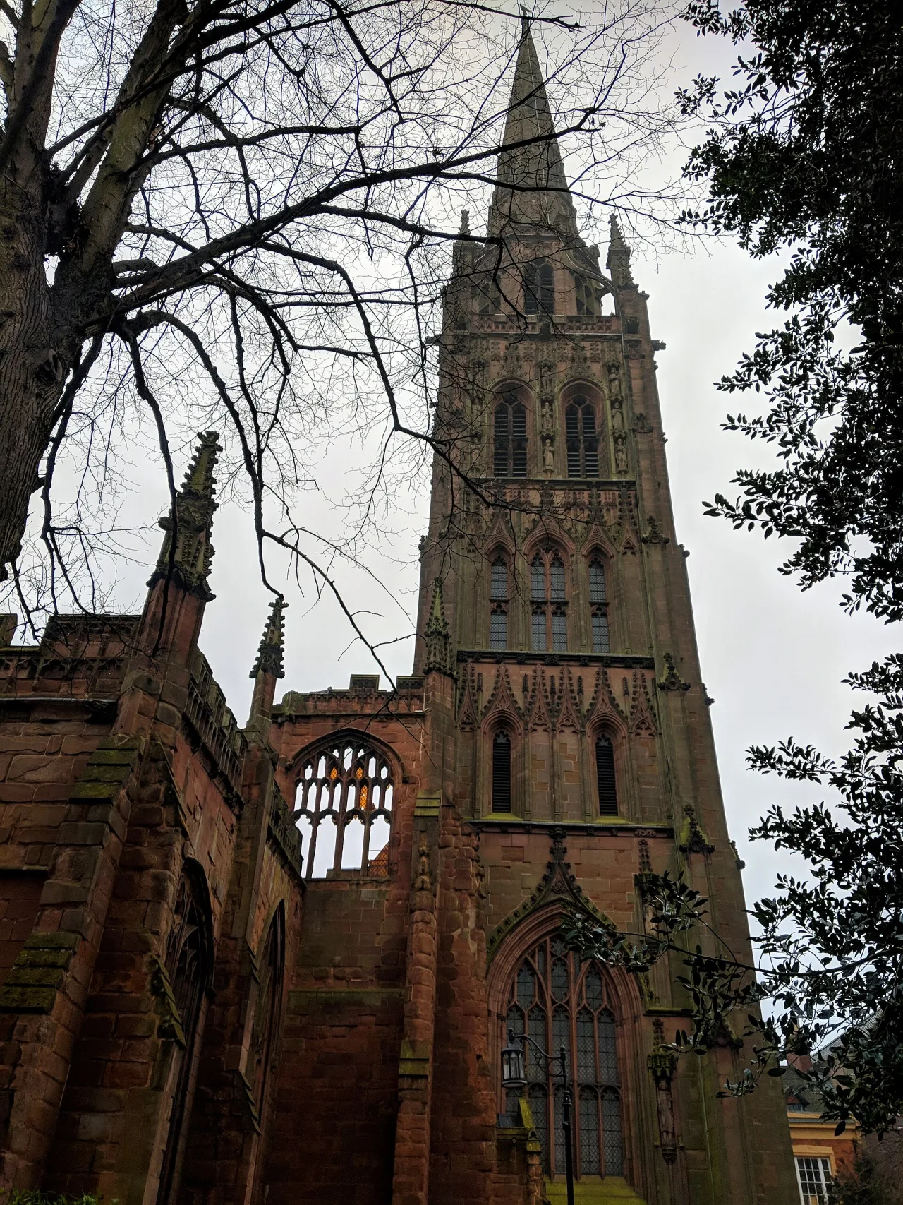 The red sandstone spire and ruined walls of the old Coventry Cathedral seen from below, framed by bare branches under a grey sky.