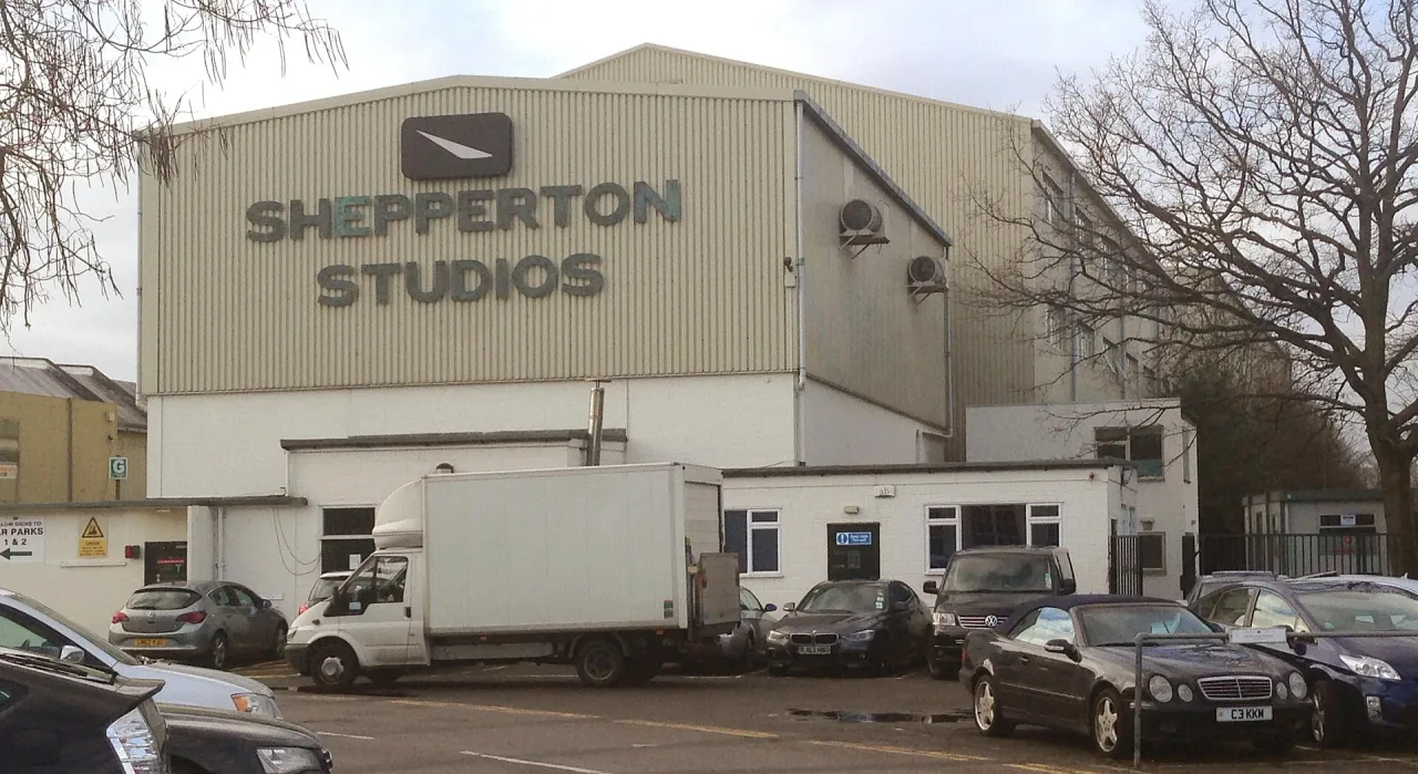 The front of a large cream-clad studio building with Shepperton Studios lettering on its wall, with parked cars and a white van in front.