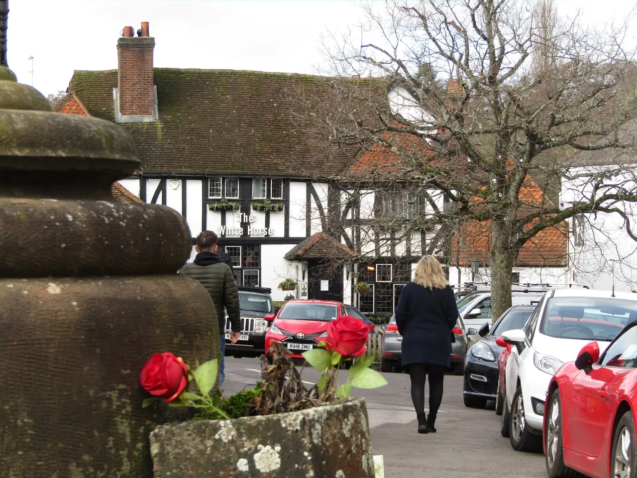The black and white timbered White Horse pub seen across the village square in Shere, with parked cars, bare trees and a stone urn with red roses in the foreground.