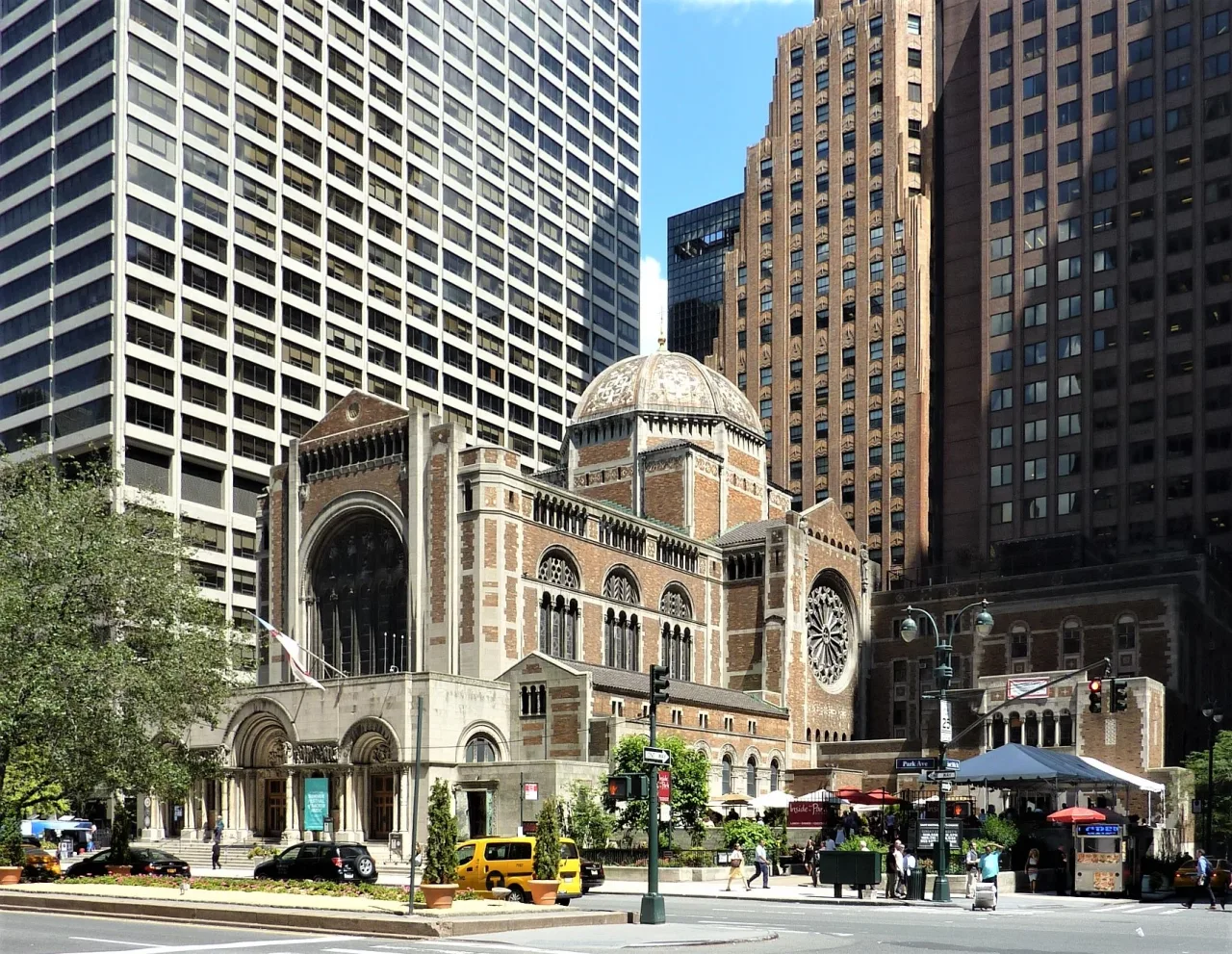 Brick and stone church with a pale domed roof and a round rose window, standing at a Manhattan street corner among tall office towers on a sunny day.