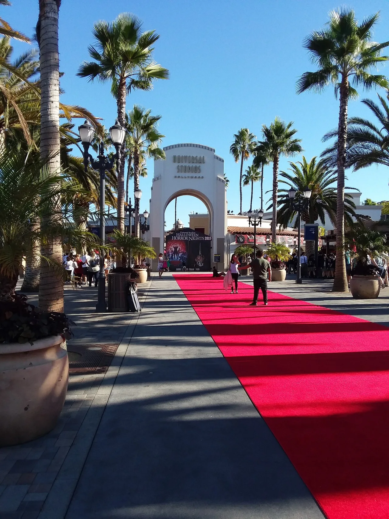 The white arched entrance of a theme park framed by palm trees and lamp posts, with a red carpet running down the right side of the walkway.