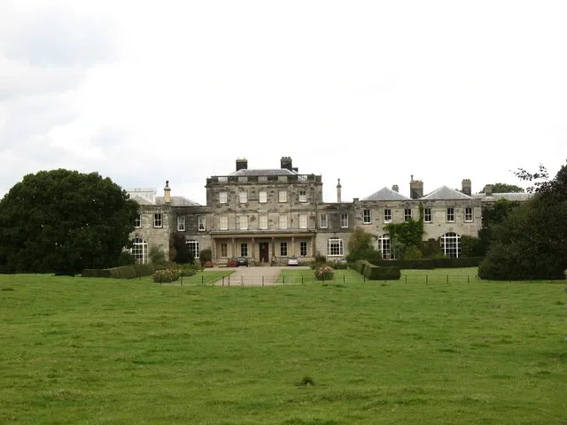 Stone-built country house with a columned central front and long wings, seen across a wide green lawn under a pale sky
