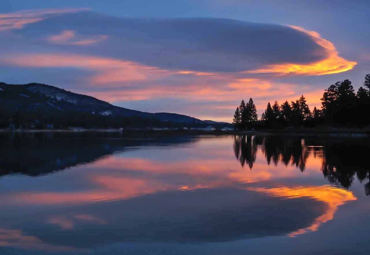 Big Bear Lake at dusk, still water mirroring an orange and purple sky, with a pine-covered shore and a mountain ridge beyond.