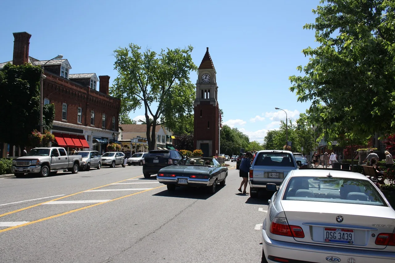 Main street of a small Ontario town on a sunny day, with a red brick clock tower, shops and parked cars.