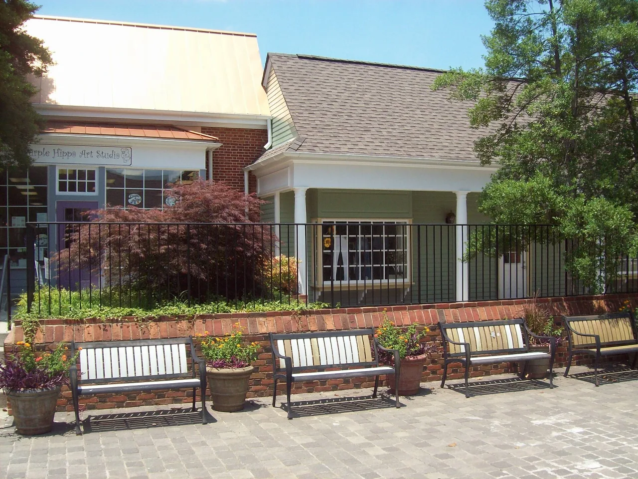 Brick-walled seating area in Dunwoody Village on a sunny day, with four park benches, flower pots and low shopfronts behind a black railing.