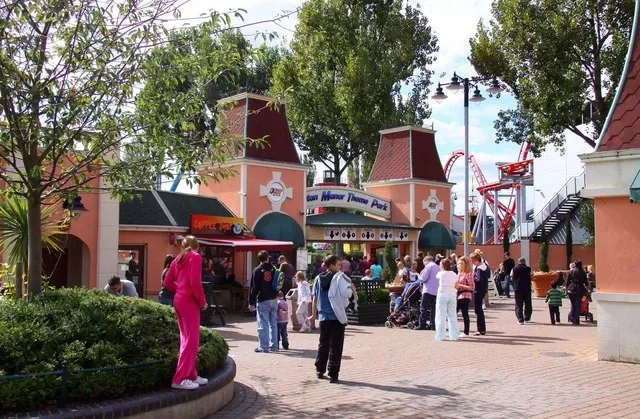 Visitors on a paved square inside a theme park, with peach-coloured buildings and a red roller coaster behind.
