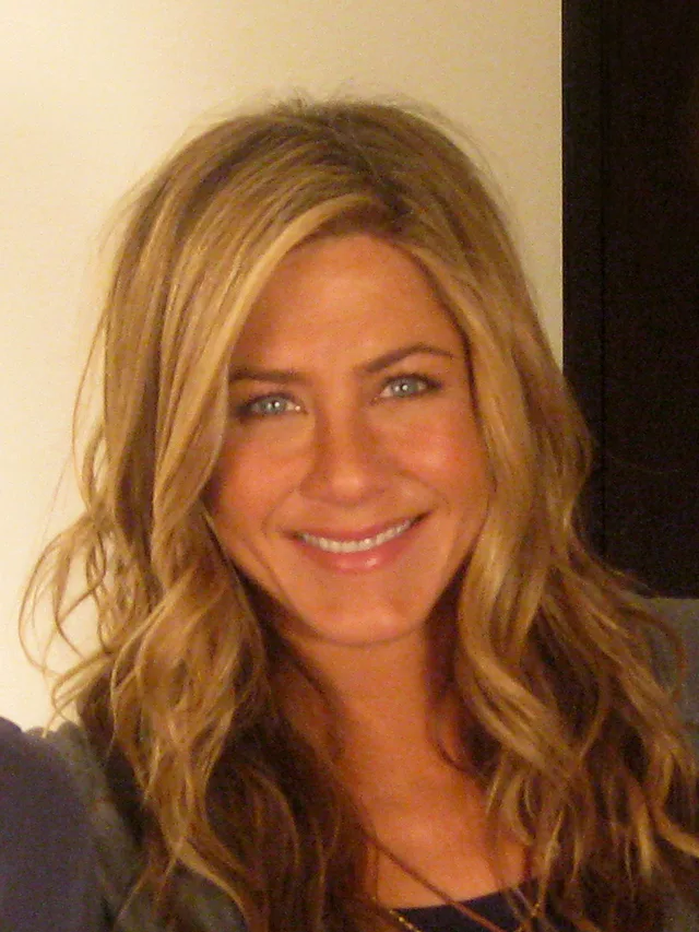 Close portrait photograph of a smiling woman with long, wavy honey-blonde hair and blue eyes, taken indoors against a cream wall.