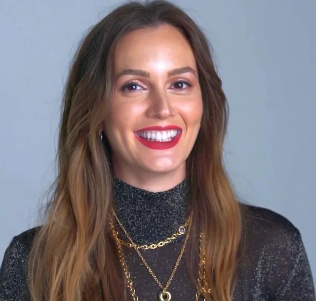 A smiling woman with long brown hair and red lipstick, in a sparkly dark high-neck top with layered gold necklaces, against a grey backdrop.