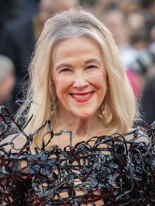 Catherine O'Hara smiling broadly, with shoulder-length blonde-grey hair and red lipstick, wearing a black sculptural top, with a blurred crowd behind her.