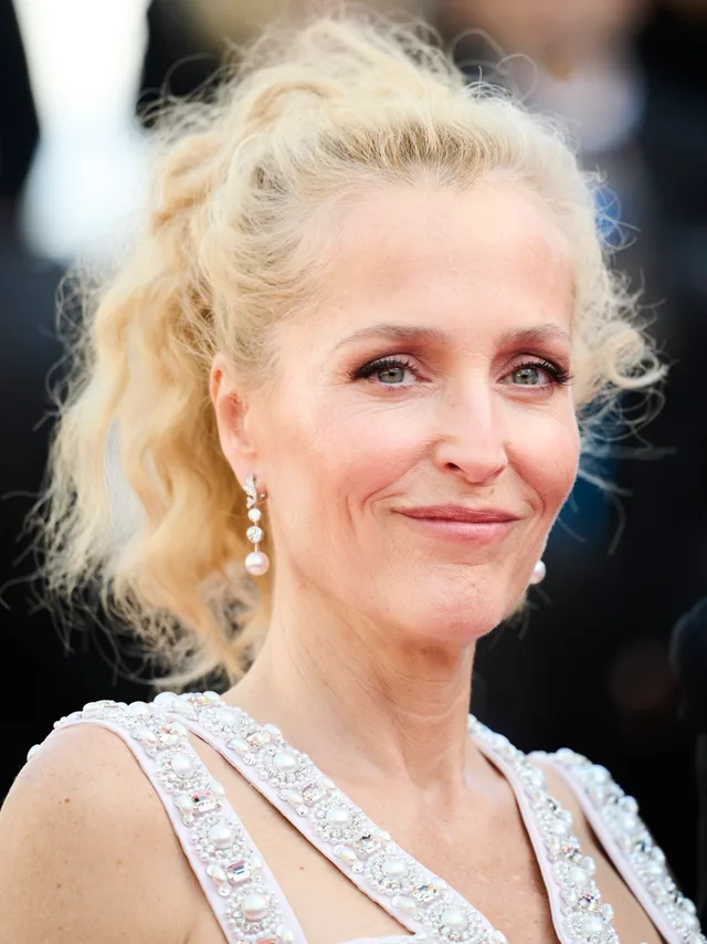 Close portrait of a woman with wavy blonde hair in a high ponytail, pearl drop earrings and a beaded pale top, smiling slightly at an evening event.