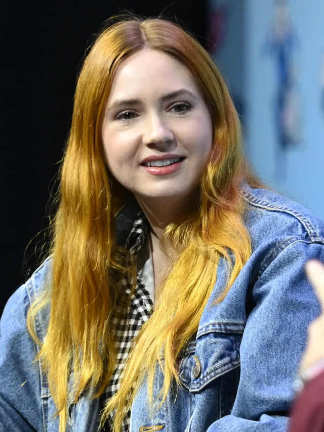 A smiling woman with long ginger hair, wearing a denim jacket over a black and white checked top, seated at a convention panel.
