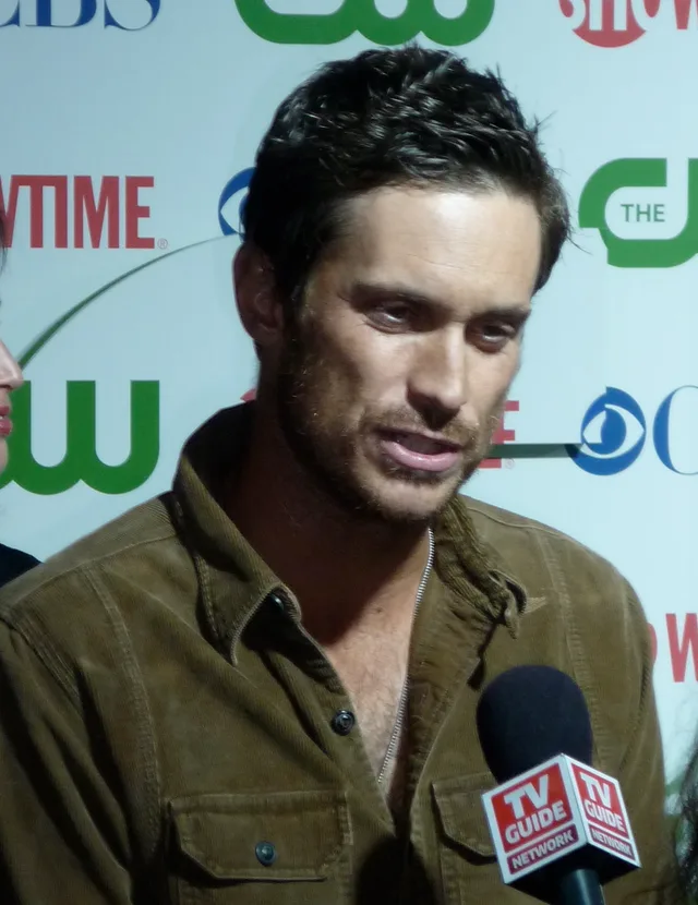 A man with dark tousled hair and stubble in a brown corduroy shirt, speaking to a microphone in front of a media backdrop.