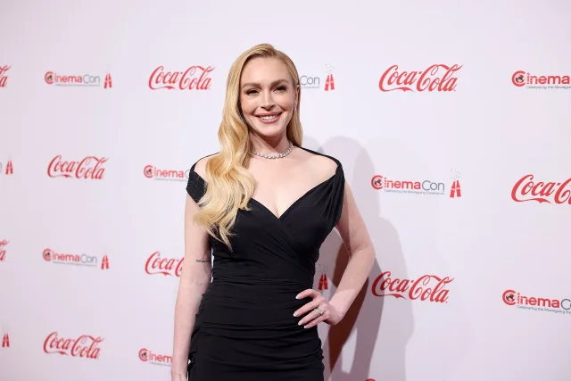 A smiling woman with long wavy blonde hair in a black wrap dress and a diamond necklace, standing with a hand on her hip on a red carpet.