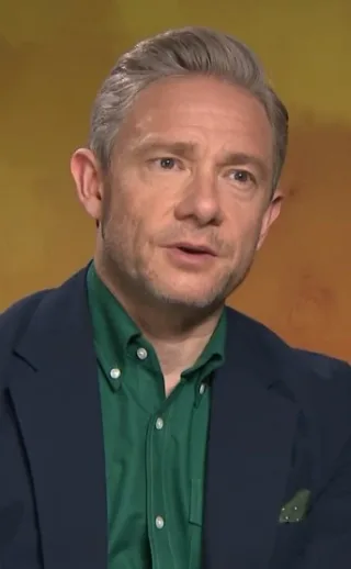 Head and shoulders photograph of Martin Freeman with swept-back grey hair, wearing a green shirt and navy jacket, speaking in front of a yellow backdrop.