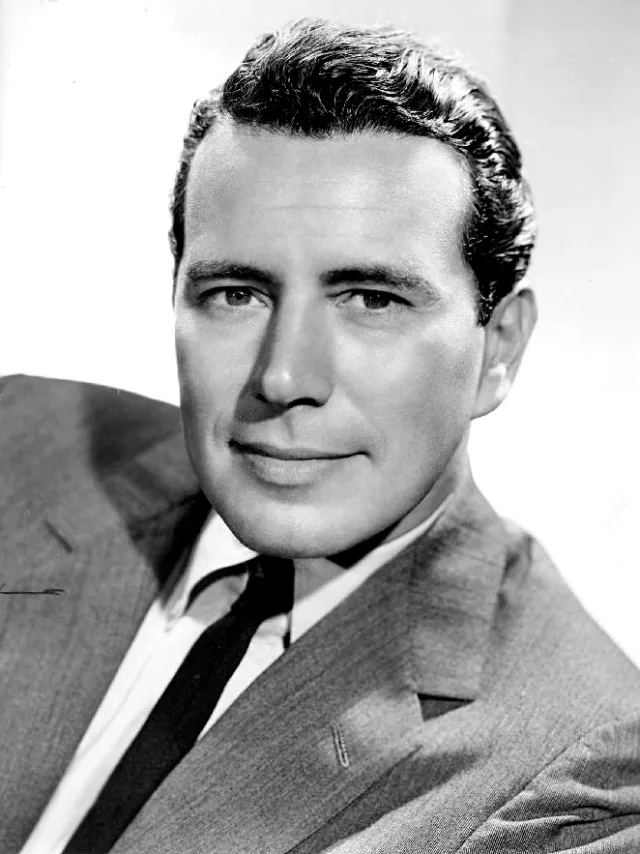 Black and white studio portrait of a smiling dark-haired man in a light suit jacket, white shirt and dark tie.