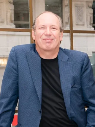 Hans Zimmer smiling slightly, in a blue checked jacket over a black top, seated by a large window.