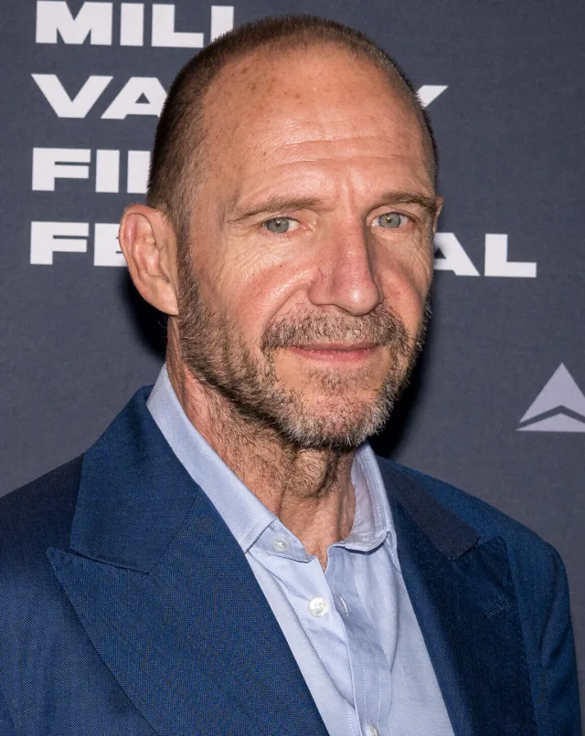 Bald man with a grey-flecked beard in a blue blazer and pale blue shirt, in front of a festival backdrop.