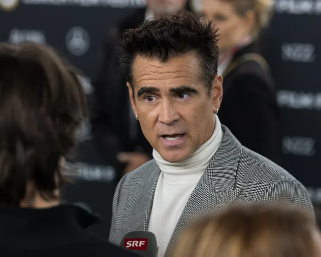 Dark-haired man in a grey checked jacket and white roll-neck speaking to a broadcast microphone at a festival press line.