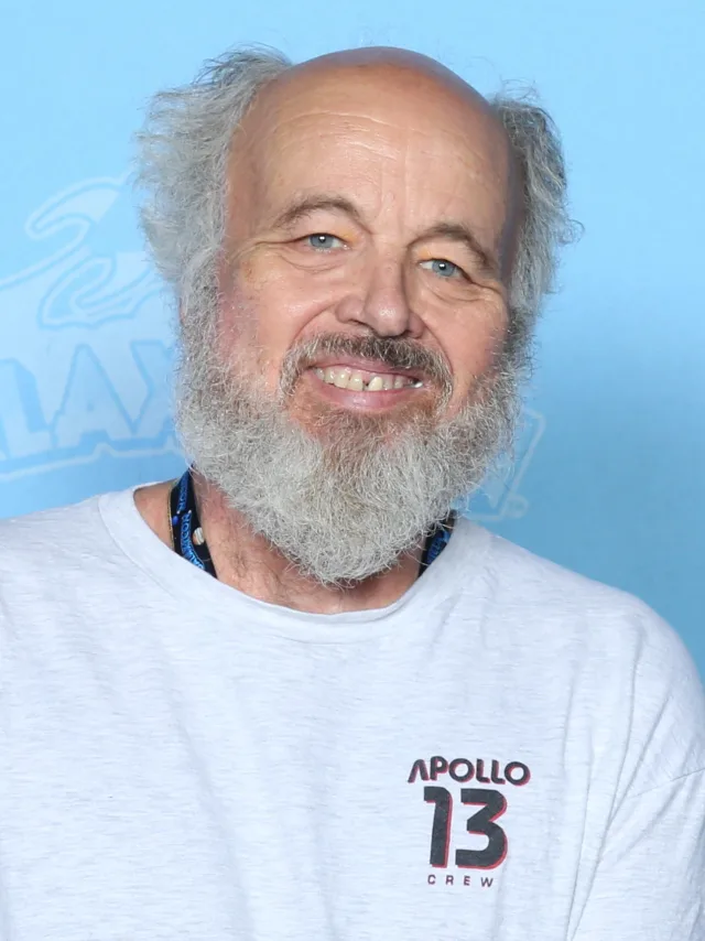 Head and shoulders photograph of a smiling older man with a full grey beard and tufts of grey hair, in a pale grey T-shirt against a light blue backdrop.