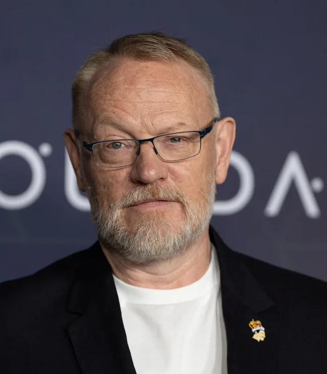 Portrait of Jared Harris with short fair hair, a grey beard and blue-framed glasses, in a dark jacket and white t-shirt.