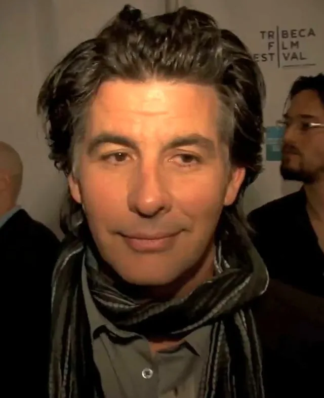 Portrait of a man with tousled dark hair and a striped scarf, photographed at a film festival press line.