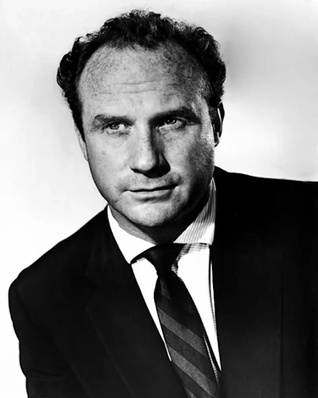 Black and white studio portrait of a man with thinning curly hair, in a dark suit and striped tie, looking off to one side.