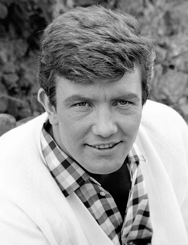 Black and white head and shoulders photograph of a smiling young man with tousled hair, wearing a checked shirt under a white cardigan, outdoors.